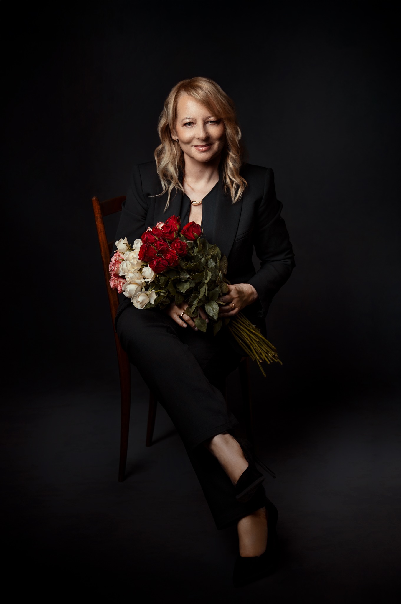 Portret kobiety w czarnym garniturze, siedzącej na krześle z bukietem róż w dłoniach na ciemnym tle. Profesjonalny wizerunek agenta nieruchomości.