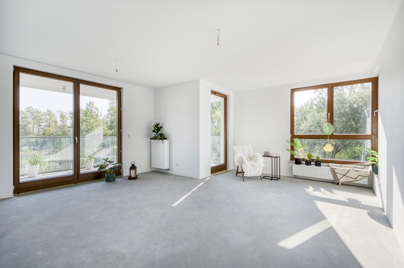 Jasne, przestronne wnętrze apartamentu z dużymi oknami i betonową podłogą. Widok na zieleń za oknem, minimalistyczny wystrój z roślinami i fotel z narzutą.