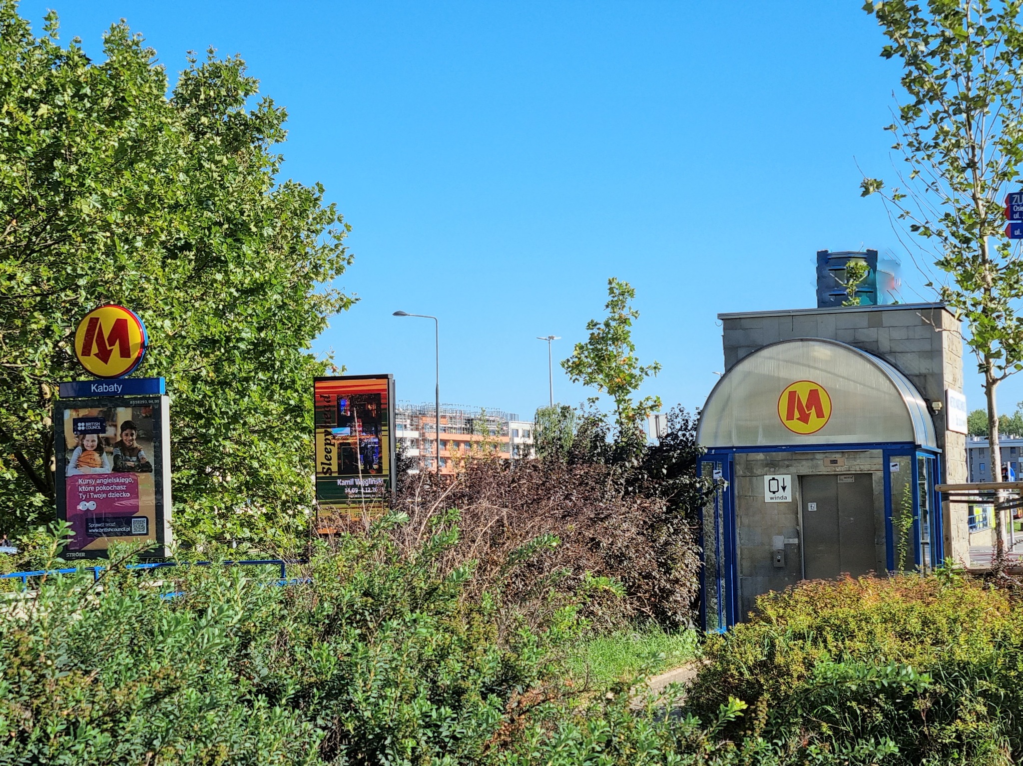 Wejście do stacji metra Kabaty w słoneczny dzień, otoczone zielenią. W tle widoczne budynki mieszkalne. Tablica reklamowa po lewej stronie.