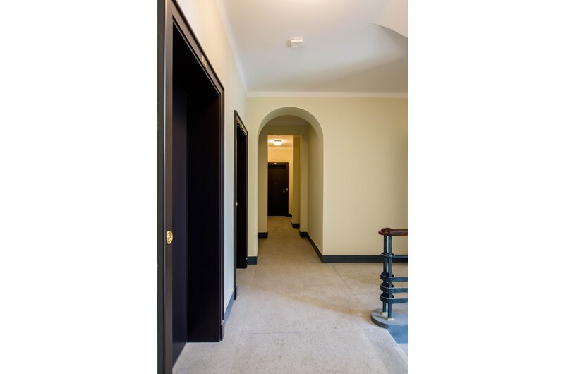 Korytarz w kamienicy z ciemnymi drzwiami i łukowym przejściem. Widoczne schody z metalową balustradą. Stonowana kolorystyka ścian i podłogi.