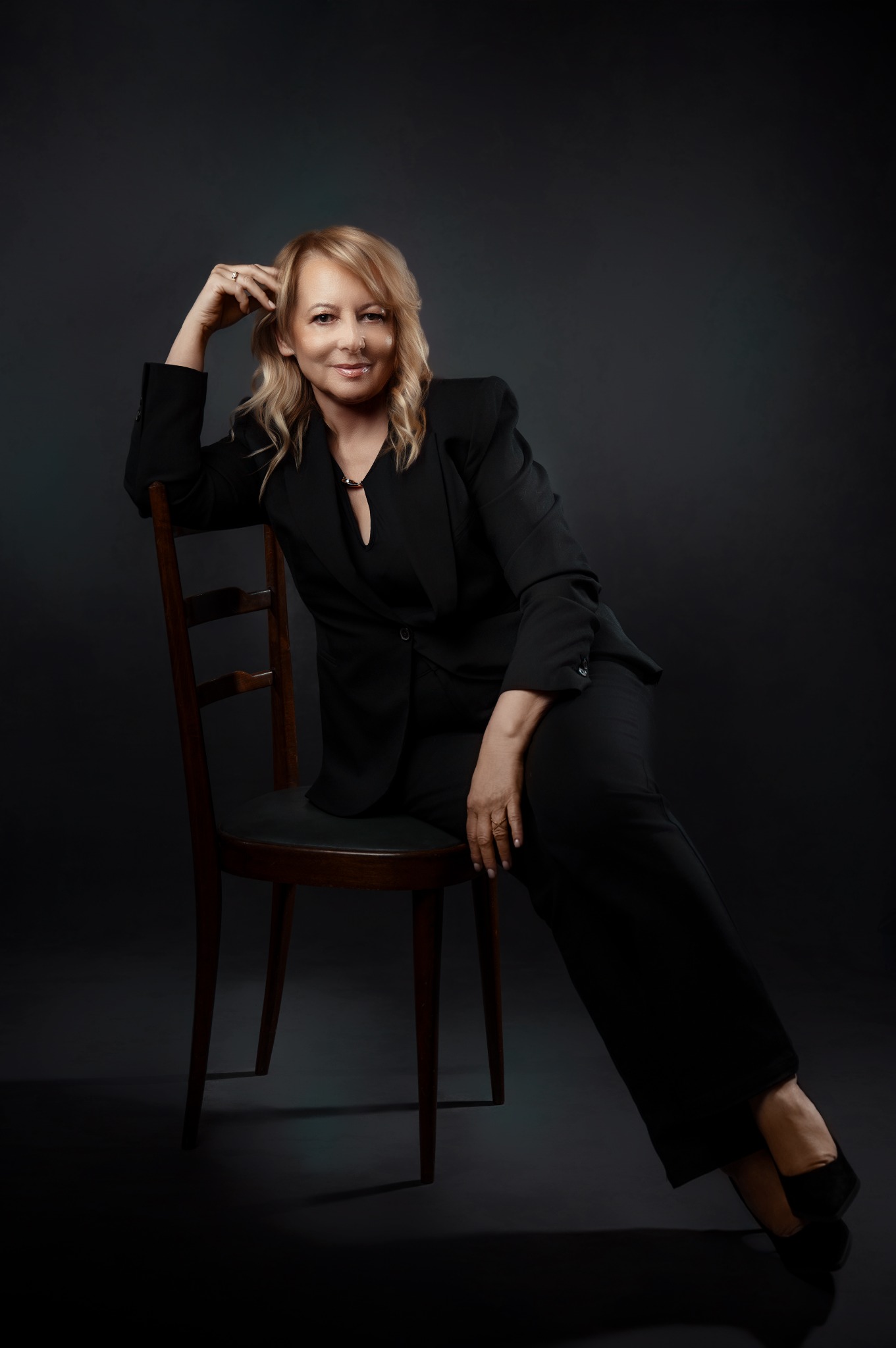 Portret kobiety w czarnym garniturze, siedzącej na drewnianym krześle na ciemnym tle. Profesjonalny wizerunek agenta nieruchomości, z delikatnym uśmiechem i naturalnym makijażem.