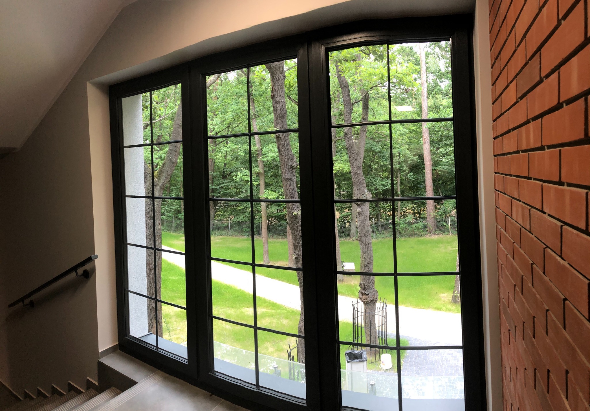 Klatka schodowa z dużym oknem z czarną ramą, widok na park z zielenią i ścieżką. Ceglana ściana po prawej stronie. Schody z szarą balustradą.