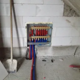 Rozdzielacz ogrzewania podłogowego wnękowy, z czerwonymi i niebieskimi rurami, zamontowany w ścianie z betonu komórkowego, widoczna łopata i fragment podłogi w trakcie prac wykończeniowych.