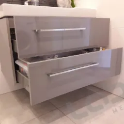Szara szafka pod umywalkę z dwiema szufladami, górna szuflada zamknięta, dolna lekko wysunięta, widoczna zawartość, nowoczesny design.