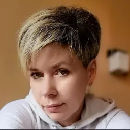 Portret kobiety z krótkimi, blond pasemkami we włosach, Szczecin, w jasnej bluzie z kapturem, na rozmytym pomarańczowym tle.