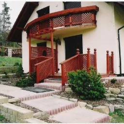 Dom z drewnianą balustradą balkonu i werandą z ażurowymi panelami. Uwagę zwraca harmonijne połączenie drewna z jasną elewacją i kamiennym obrzeżem ścieżki.