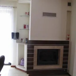 Nowoczesny kominek z obudową z ciemnego drewna i beżowego kamienia, wbudowany w ścianę. Po lewej stronie półki z dekoracjami i zasłonięte okno.