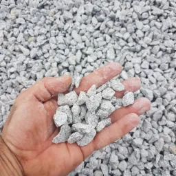 Otwarta dłoń trzymająca garść drobnego, szarego kruszywa na tle większej ilości tego samego materiału.