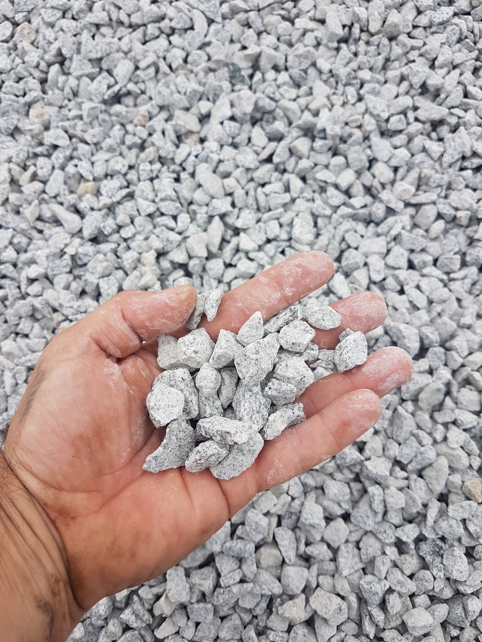 Otwarta dłoń trzymająca garść drobnego, szarego kruszywa na tle większej ilości tego samego materiału.