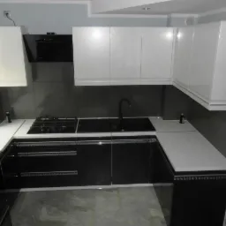 Nowoczesna kuchnia w czerni i bieli, z błyszczącymi frontami szafek i jasnym blatem. Widoczny zlew, płyta gazowa i okap. Uwagę zwraca minimalistyczny design i kontrast kolorów.