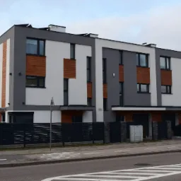 Nowoczesny trzypiętrowy budynek mieszkalny z elementami drewnianej elewacji i balkonem, widoczny z ulicy z oznakowanym przejściem dla pieszych i rowerzystów.