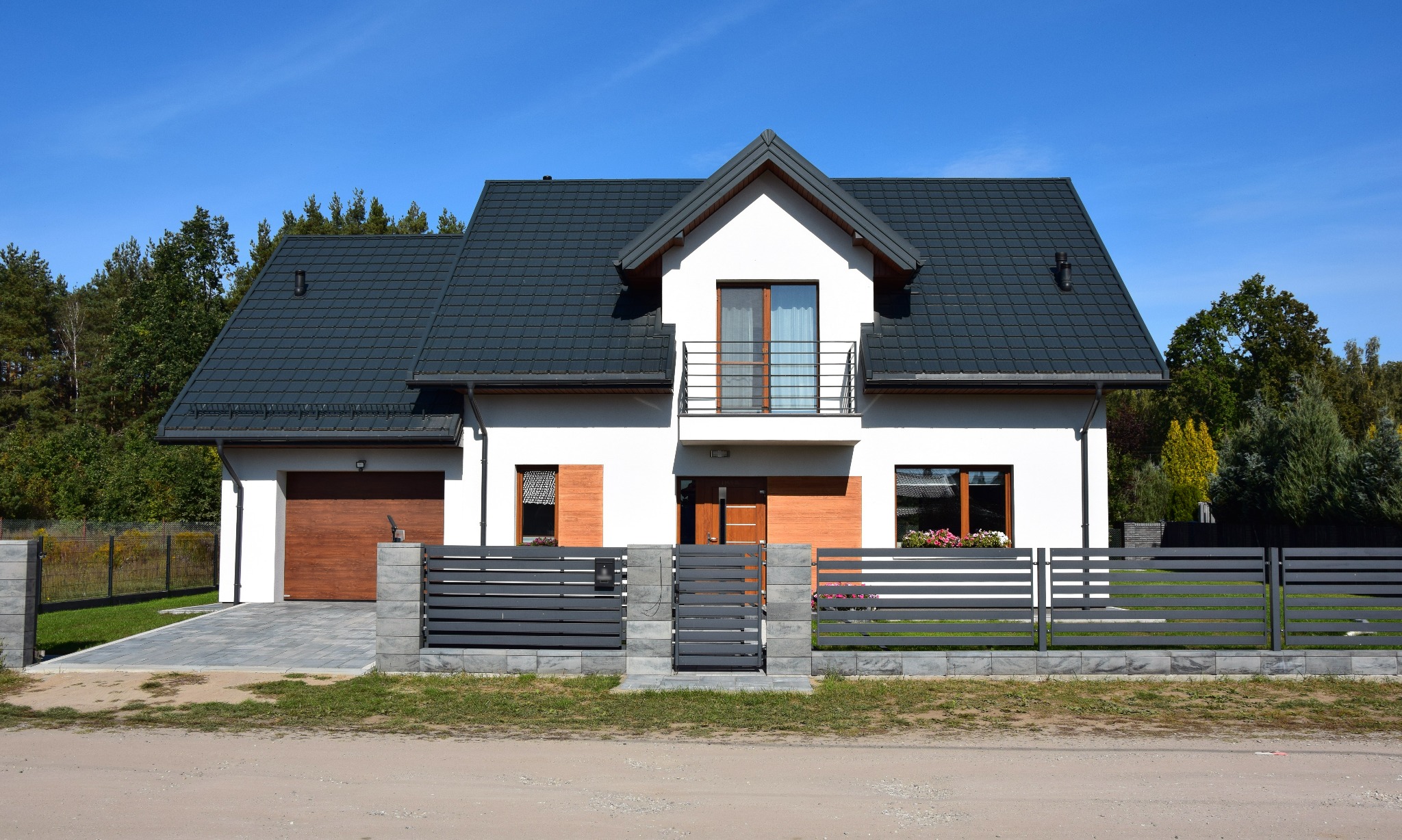 Nowoczesny, dwukondygnacyjny dom z ciemną dachówką i drewnianymi akcentami, otoczony szarym ogrodzeniem panelowym i zielenią. Błękitne niebo w tle.