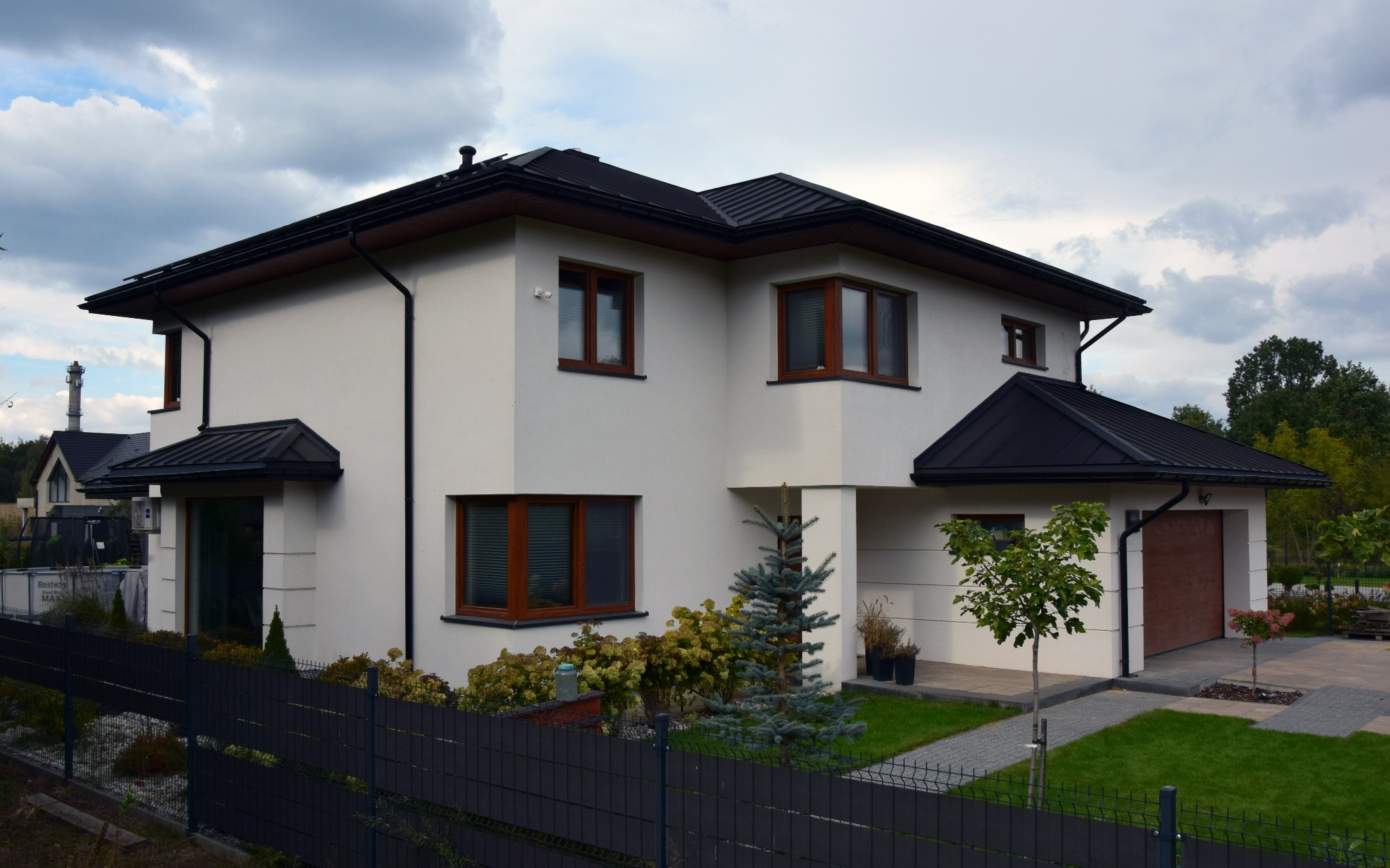 Dwukondygnacyjny dom z białymi ścianami, brązowymi oknami i czarnym dachem, otoczony zielenią i ogrodzeniem. Widok z zewnątrz, w tle pochmurne niebo.