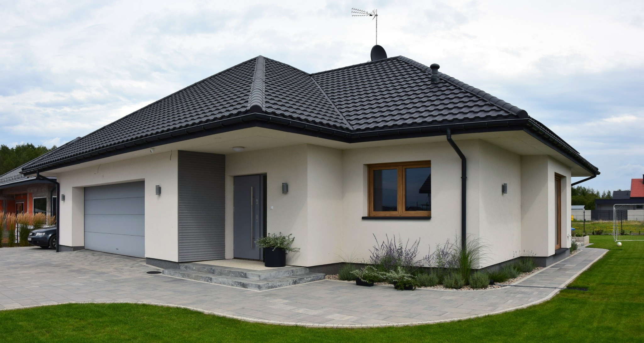 Parterowy dom z garażem i ciemną dachówką, jasna elewacja z szarymi akcentami. Uwagę zwraca starannie zagospodarowany teren wokół budynku i nowoczesna bryła.