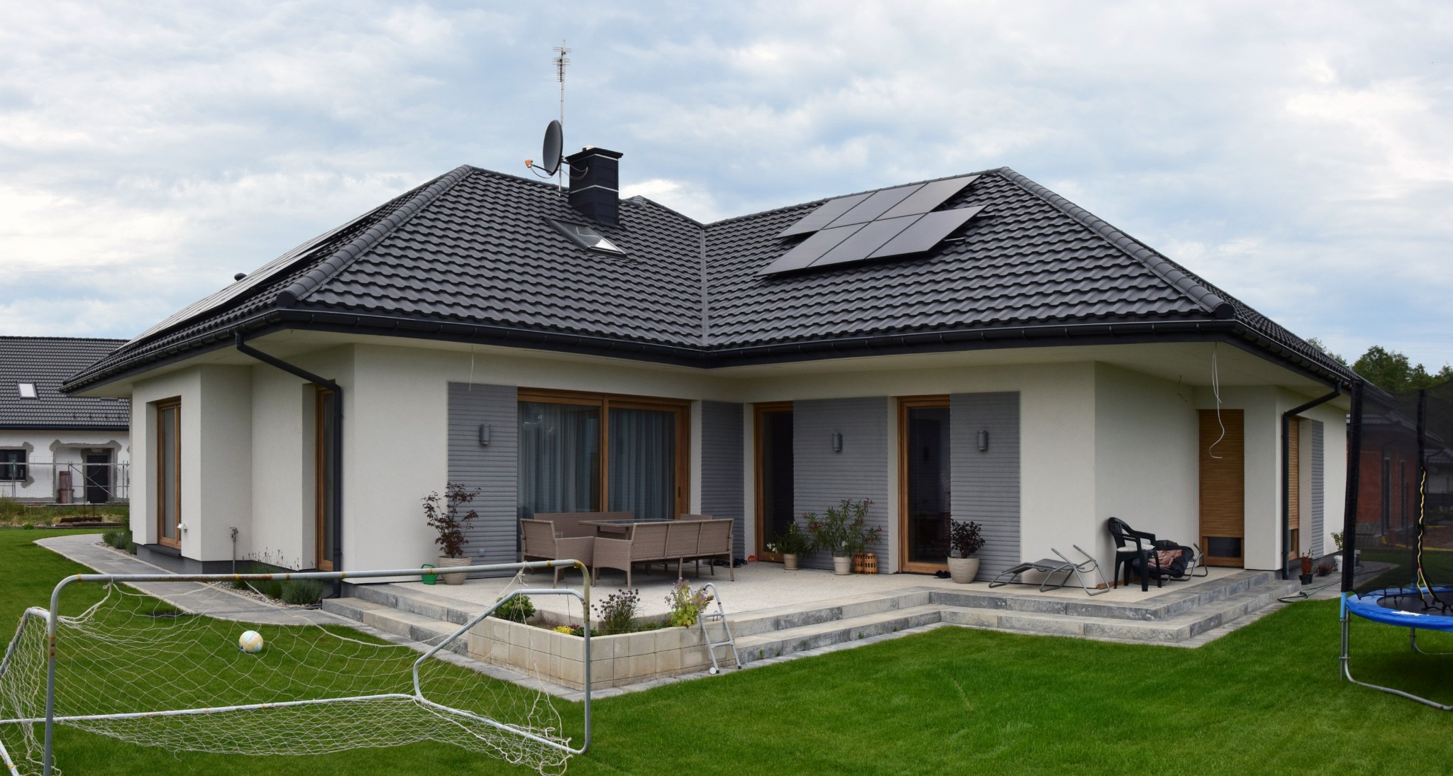 Nowoczesny dom jednorodzinny z panelami słonecznymi na dachu i tarasem z meblami ogrodowymi, otoczony zielonym trawnikiem z bramką piłkarską.