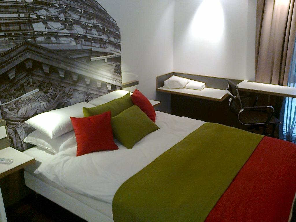 Pokój hotelowy z łóżkiem, biurkiem i fototapetą przedstawiającą budowlę. Czerwono-zielona kolorystyka pościeli. Minimalistyczny wystrój wnętrza.