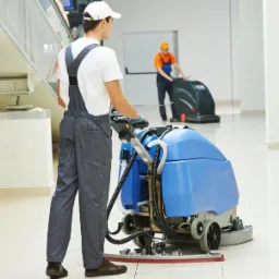 Pracownik w kombinezonie obsługuje automat do mycia podłóg w jasnym, przeszklonym biurze. W tle inny pracownik z podobnym urządzeniem.