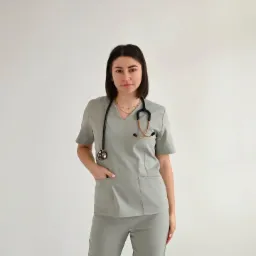 Kobieta w szarym uniformie medycznym ze stetoskopem na szyi, pozuje na jasnym tle. Widoczna kieszeń na bluzie i spodnie, całość w neutralnym odcieniu szarości.