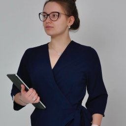 KEIA Paulina Krzeszewska - Kobieta w granatowej sukience z kopertowym dekoltem, trzymająca tablet. Elegancki strój, upięte włosy, okulary i delikatna biżuteria. Jasne tło.