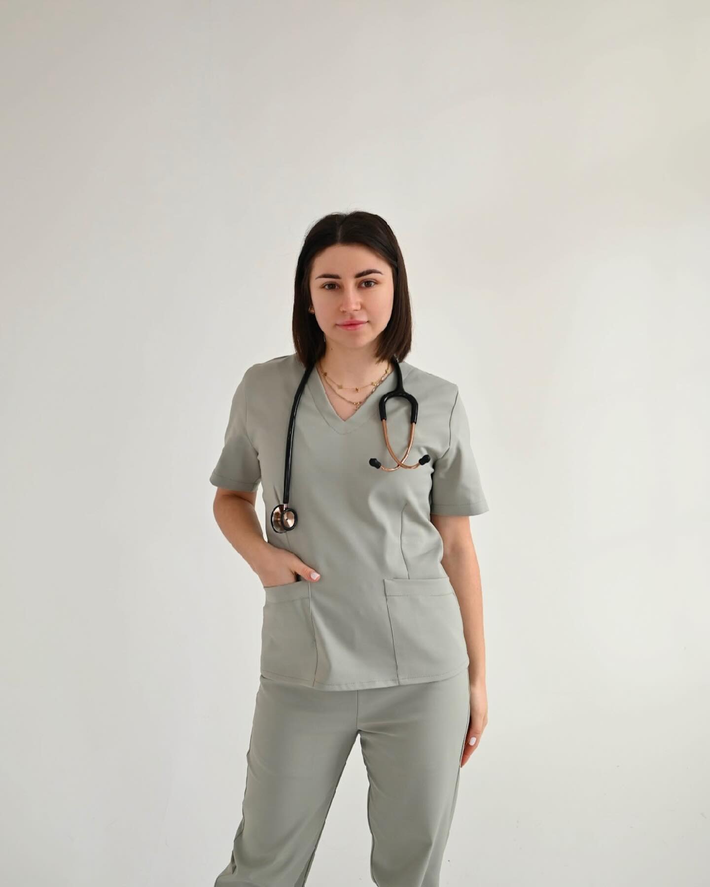 Kobieta w szarym uniformie medycznym ze stetoskopem na szyi, pozuje na jasnym tle. Widoczna kieszeń na bluzie i spodnie, całość w neutralnym odcieniu szarości.
