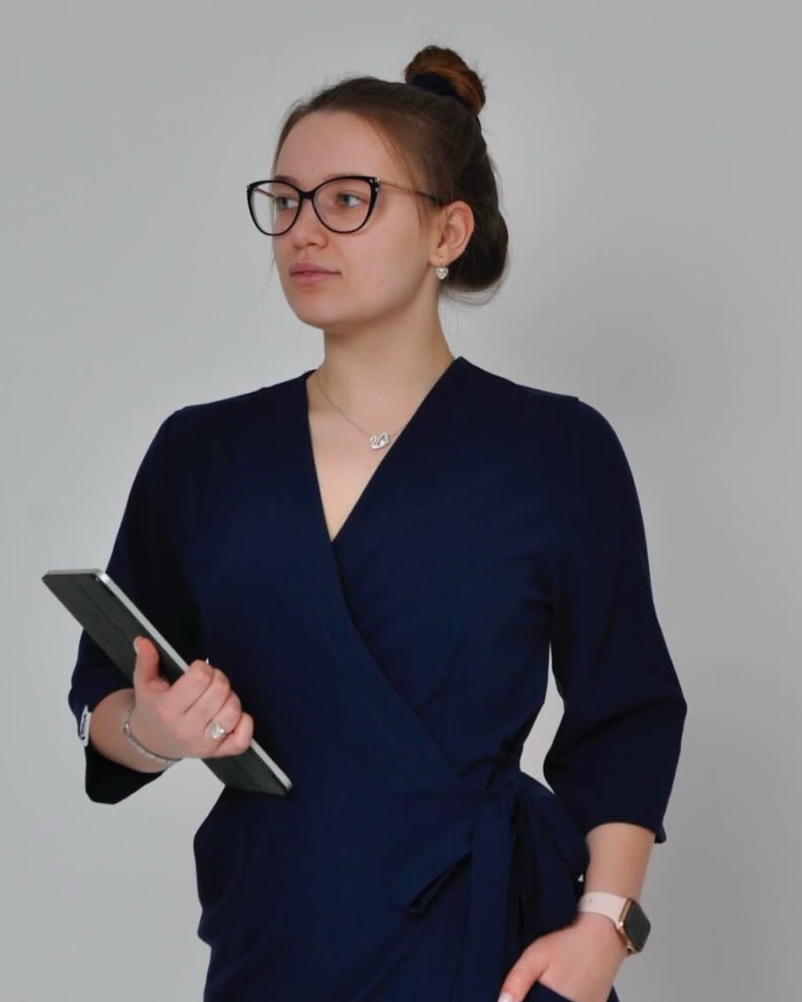 Kobieta w granatowej sukience z kopertowym dekoltem, trzymająca tablet. Elegancki strój, upięte włosy, okulary i delikatna biżuteria. Jasne tło.