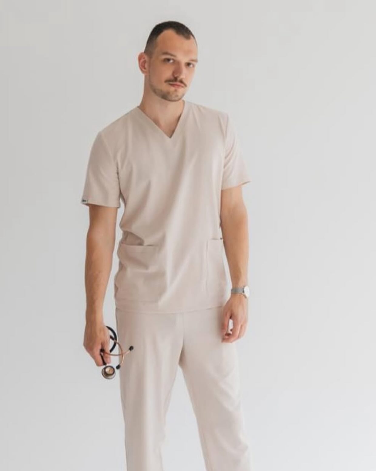 Mężczyzna w beżowym komplecie odzieży roboczej medycznej, trzymający stetoskop. Widoczny zegarek na ręku i gładkie tło, sugerujące studyjną sesję.