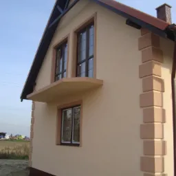 Elewacja domu jednorodzinnego po tynkowaniu i malowaniu, z dekoracyjnymi elementami narożnymi i niewielkim balkonem pod oknami na poddaszu. Jasna kolorystyka.