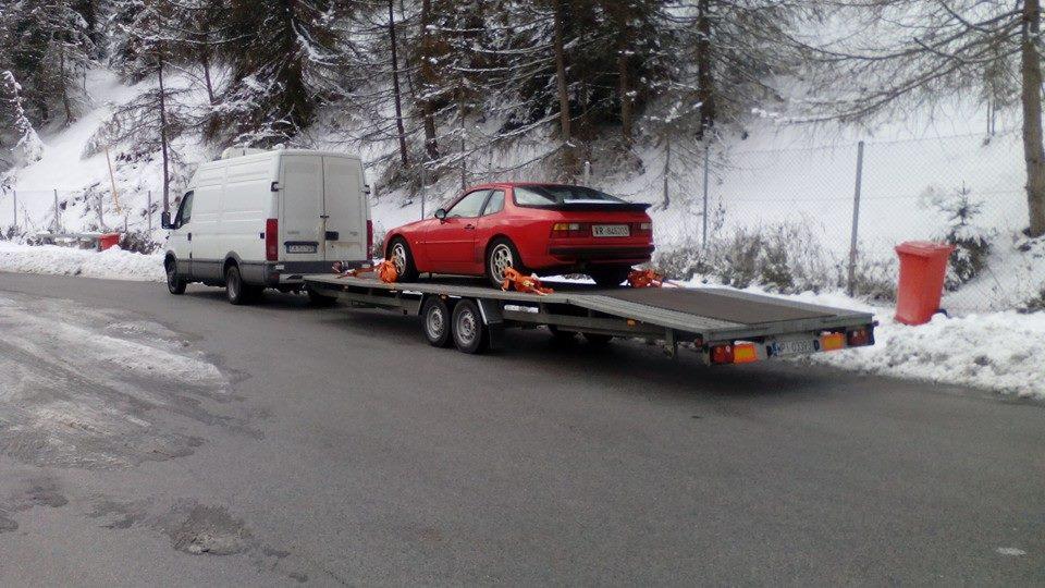 Biały van ciągnie lawetę z czerwonym Porsche 944 na tle ośnieżonego lasu.