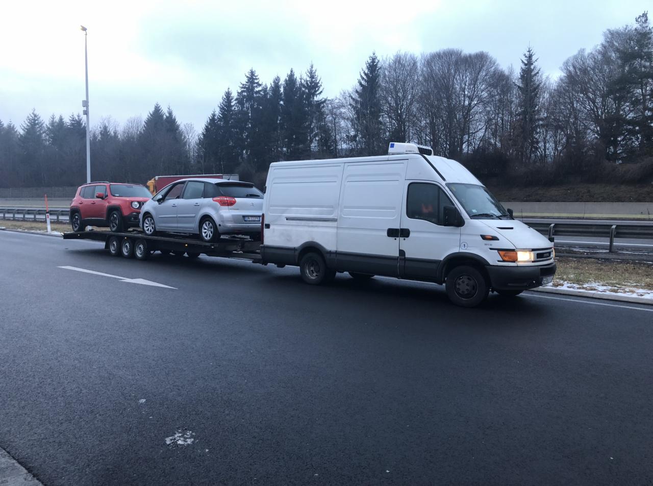 Biała laweta Iveco z agregatem chłodniczym przewozi dwa samochody osobowe: czerwonego Jeepa Renegade i srebrnego Citroena C4 na autostradzie w pochmurny dzień.