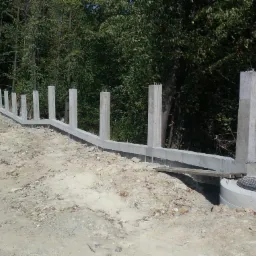 Wznoszenie betonowego muru oporowego z wystającymi prętami zbrojeniowymi, widok z boku na tle drzew. Paleta materiałów budowlanych z lewej strony kadru.