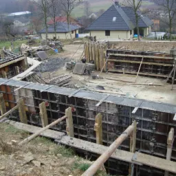 Szczegółowe ujęcie szalunków fundamentowych na placu budowy z widocznymi elementami drewnianymi i metalowymi, w tle zabudowa jednorodzinna i drzewa.