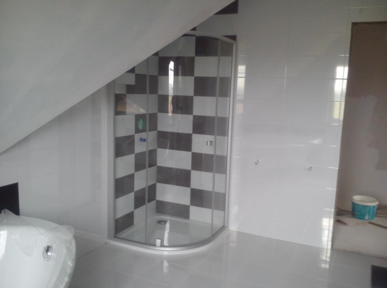 Nowoczesna kabina prysznicowa w łazience na poddaszu, wyłożona biało-szarą szachownicą. Widoczne białe płytki na ścianach i podłodze, oraz fragment wanny pod folią.