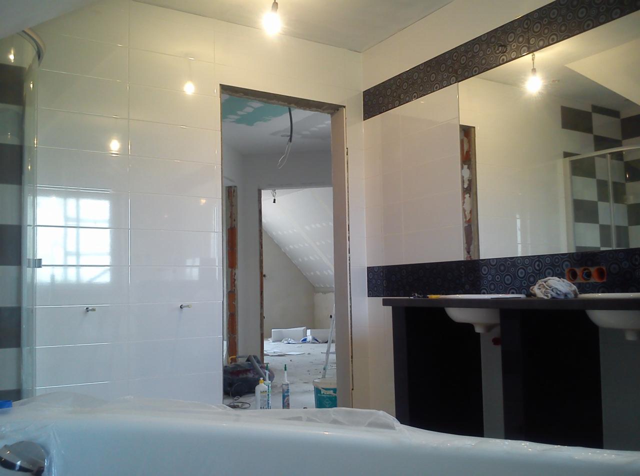 Wykończona łazienka z białą glazurą, dekoracyjnym pasem na ścianie z lustrem. W tle widoczne pomieszczenie w trakcie prac wykończeniowych na poddaszu.