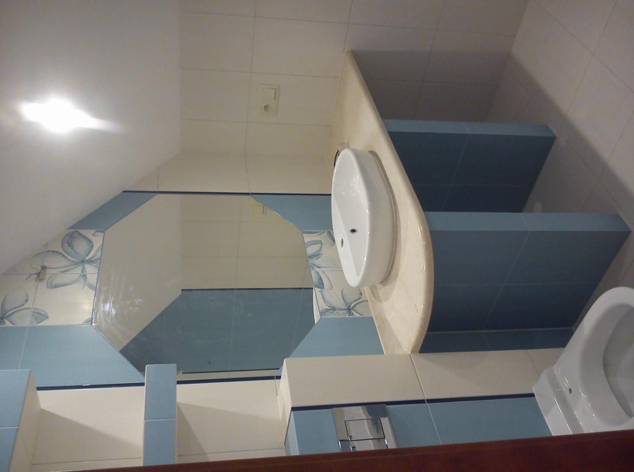 Wykończona łazienka na poddaszu: biała umywalka na blacie z marmuru, toaleta i lustro z niebieskimi akcentami. Płytki z motywem kwiatowym. Oświetlenie sufitowe.