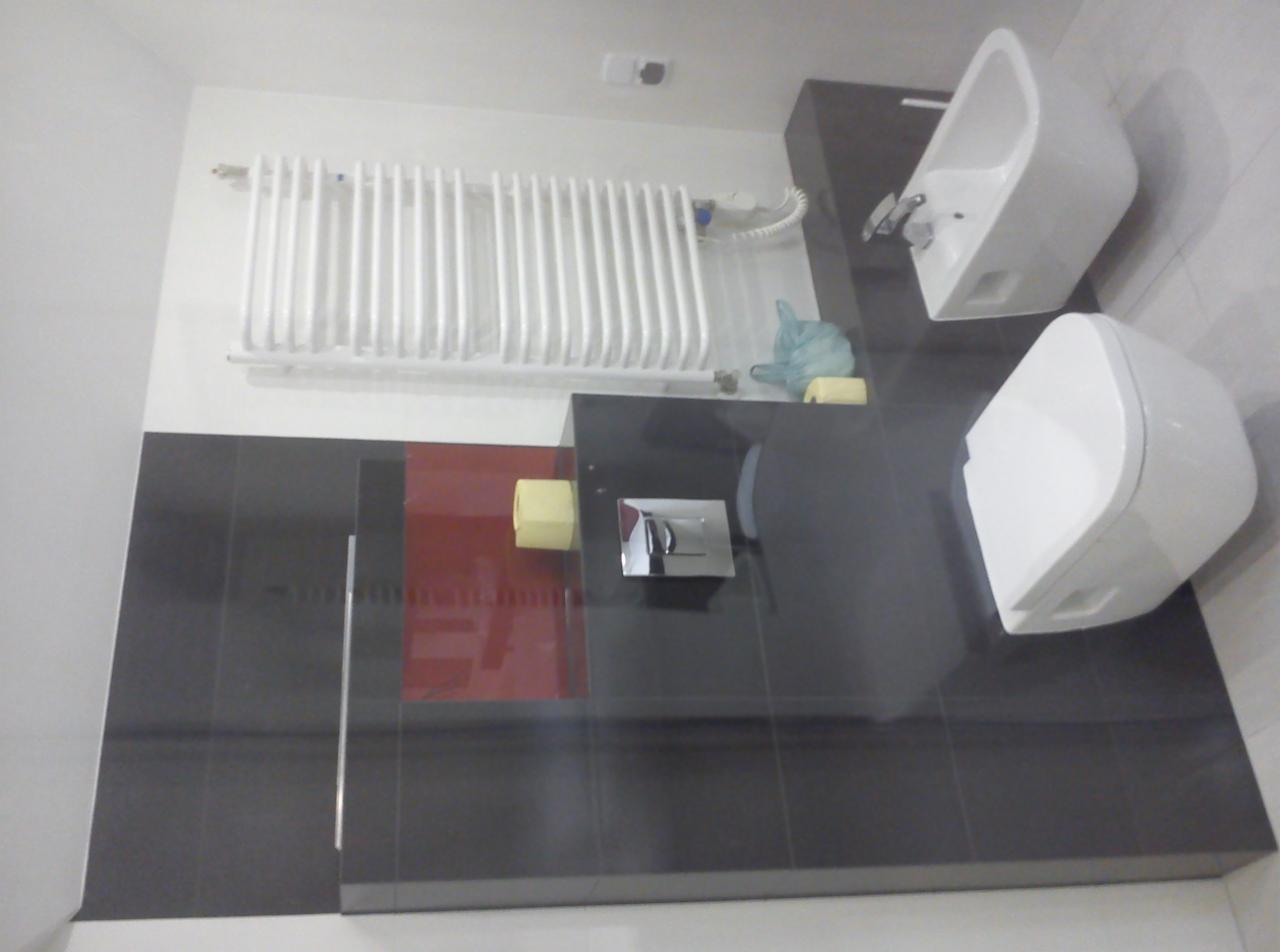 Wyremontowana łazienka z czarnymi płytkami, białą toaletą i bidetem. Na ścianie biały grzejnik. Czerwony akcent na ścianie dodaje charakteru.