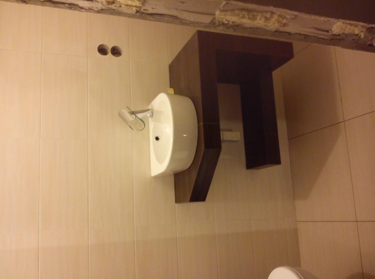 Biała umywalka na brązowej szafce w łazience z beżowymi płytkami. Widoczne dwa otwory w ścianie. Ujęcie z dołu, lekko obrócone.