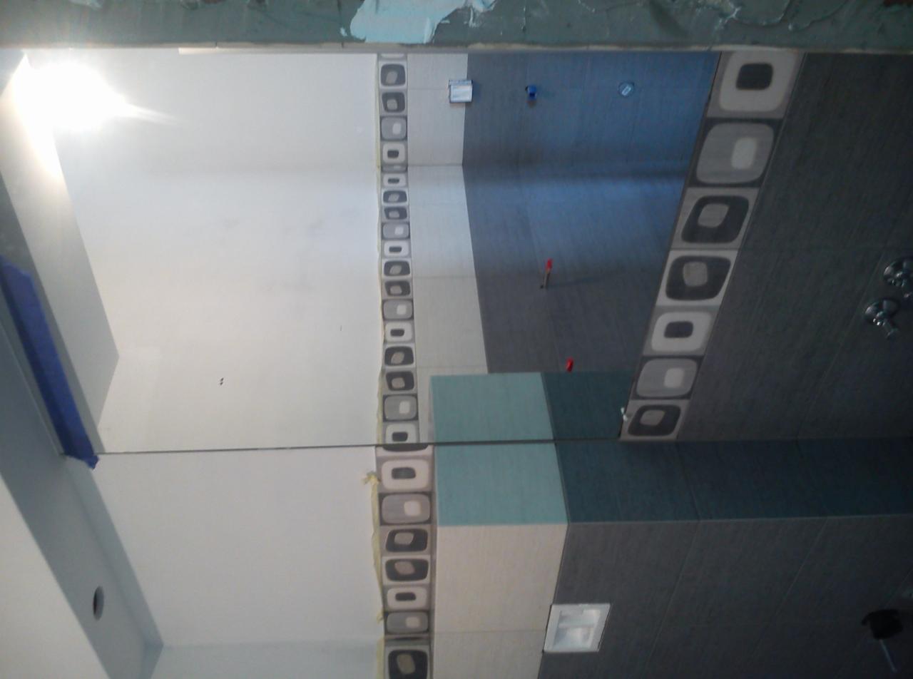 Ściana łazienki w trakcie wykańczania: ułożone płytki w odcieniach szarości, bieli i turkusu z dekoracyjnym pasem z motywem kwadratów. Widoczne elementy montażowe, surowe wykończenie.