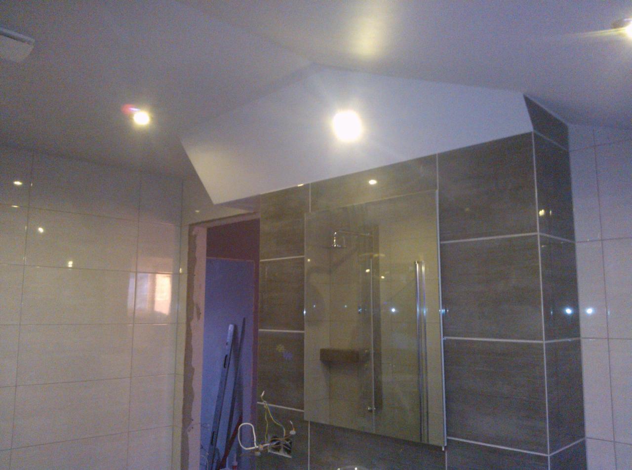 Wykończona łazienka z szarymi i białymi płytkami, kabina prysznicowa ze szklanymi drzwiami, podwieszany sufit z oświetleniem punktowym. Widoczne narzędzia budowlane.