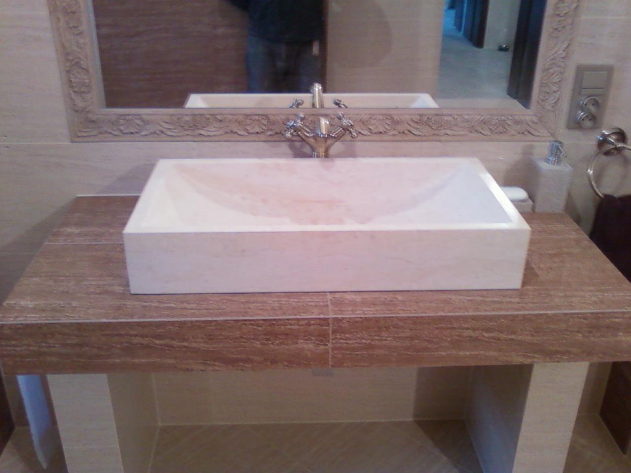 Beżowa, prostokątna umywalka z marmuru, z ozdobną armaturą, osadzona na blacie z płytek imitujących drewno. W tle lustro z rzeźbioną ramą.