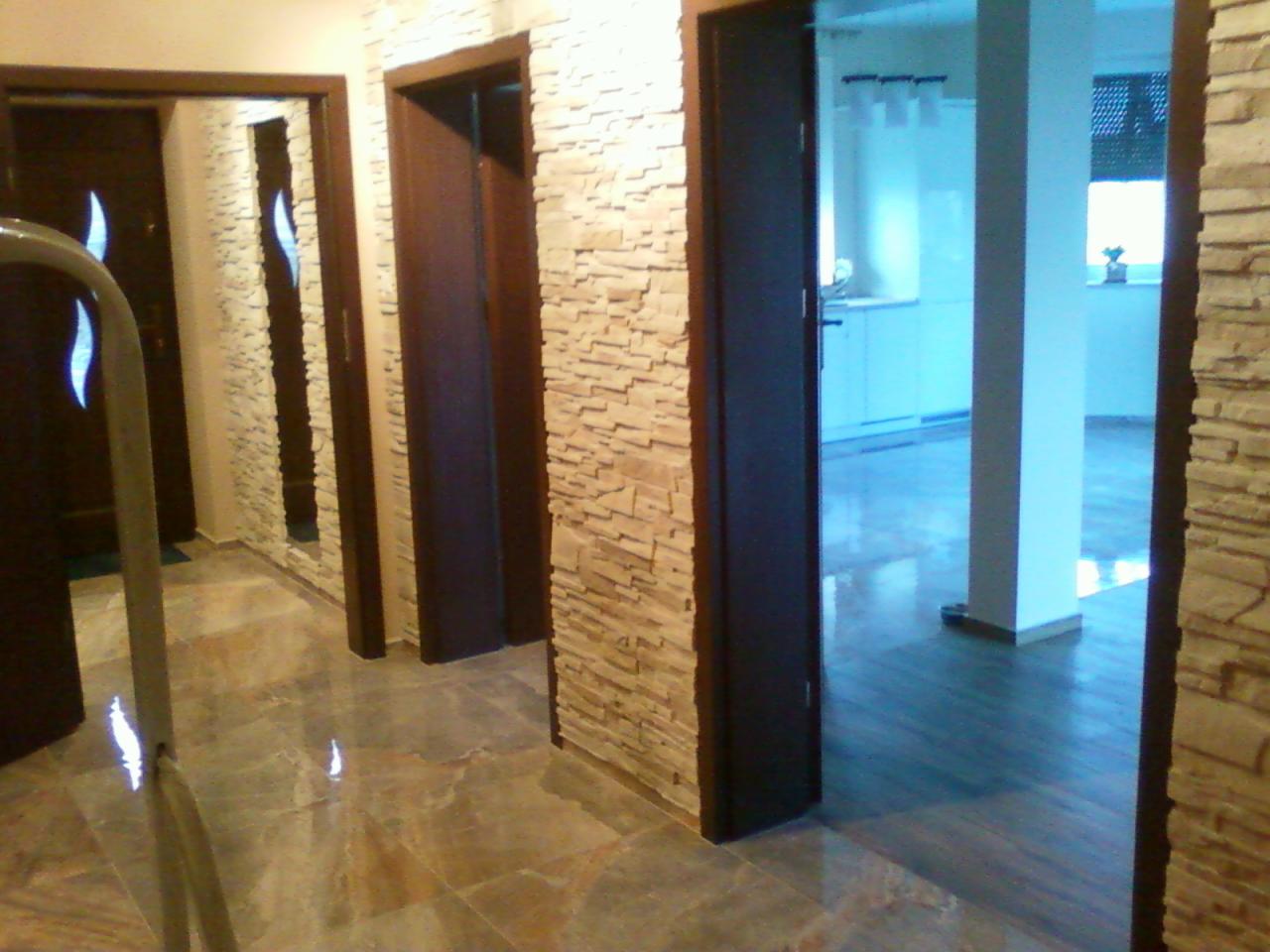 Korytarz z kamienną okładziną ścian, ciemnymi drzwiami i marmurową podłogą. W tle widoczna kuchnia z białymi meblami. Oświetlenie sufitowe.