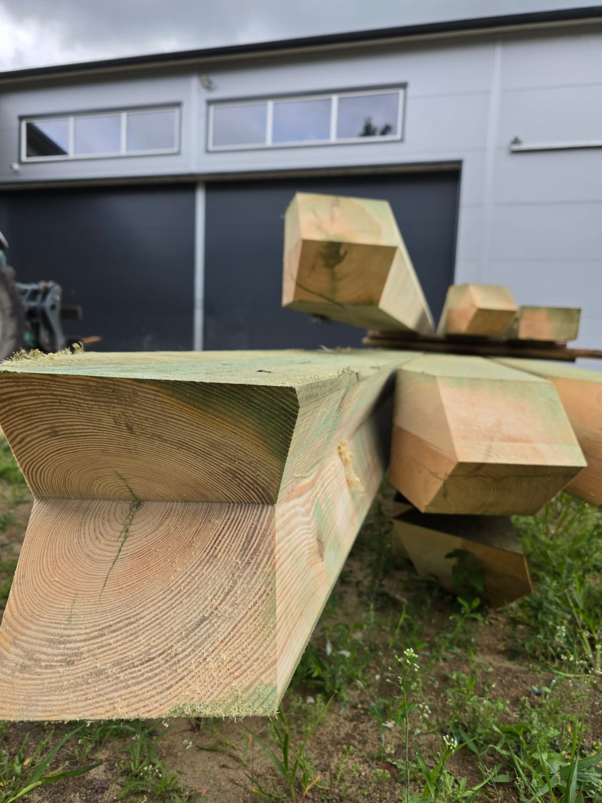 Świeżo obrobione, impregnowane drewno konstrukcyjne z widocznymi słojami, ułożone na trawie przed budynkiem z szarą elewacją i czarną bramą.