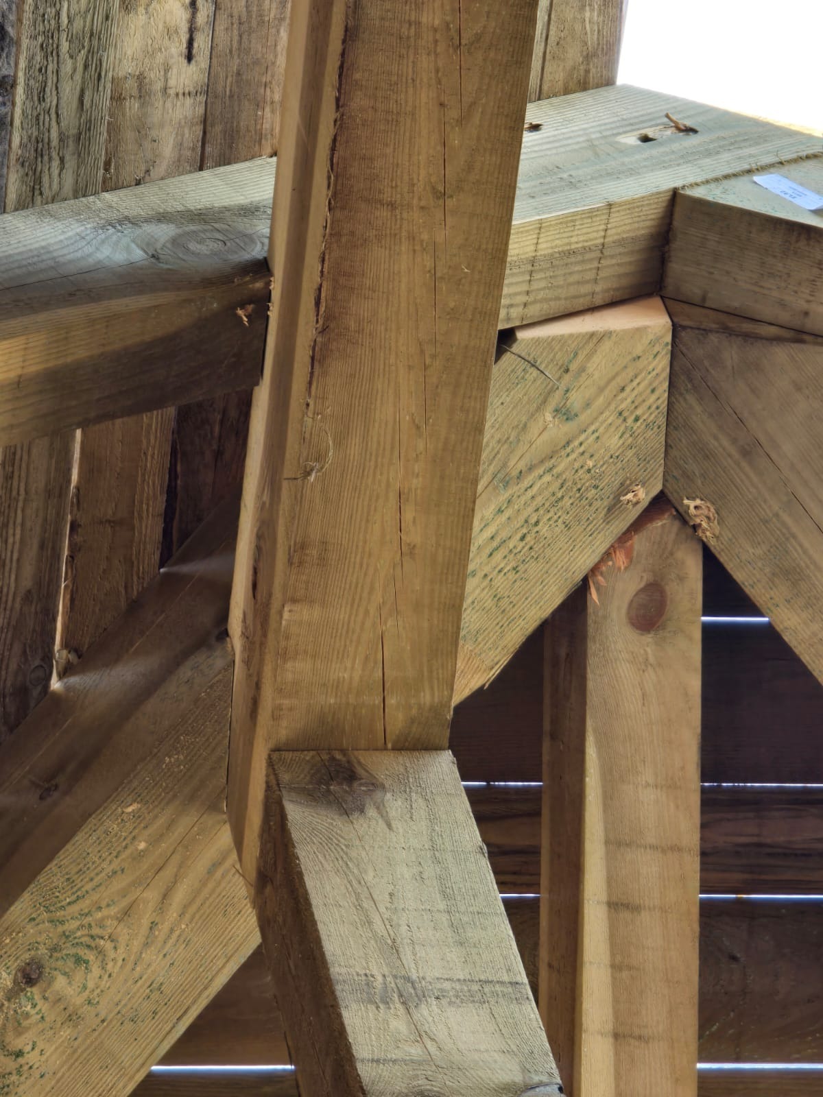 Konstrukcja drewniana z belek i słupów, widok z dołu, ukazujący połączenia elementów. Widoczne ślady obróbki drewna i impregnacji. Jasne, naturalne oświetlenie.