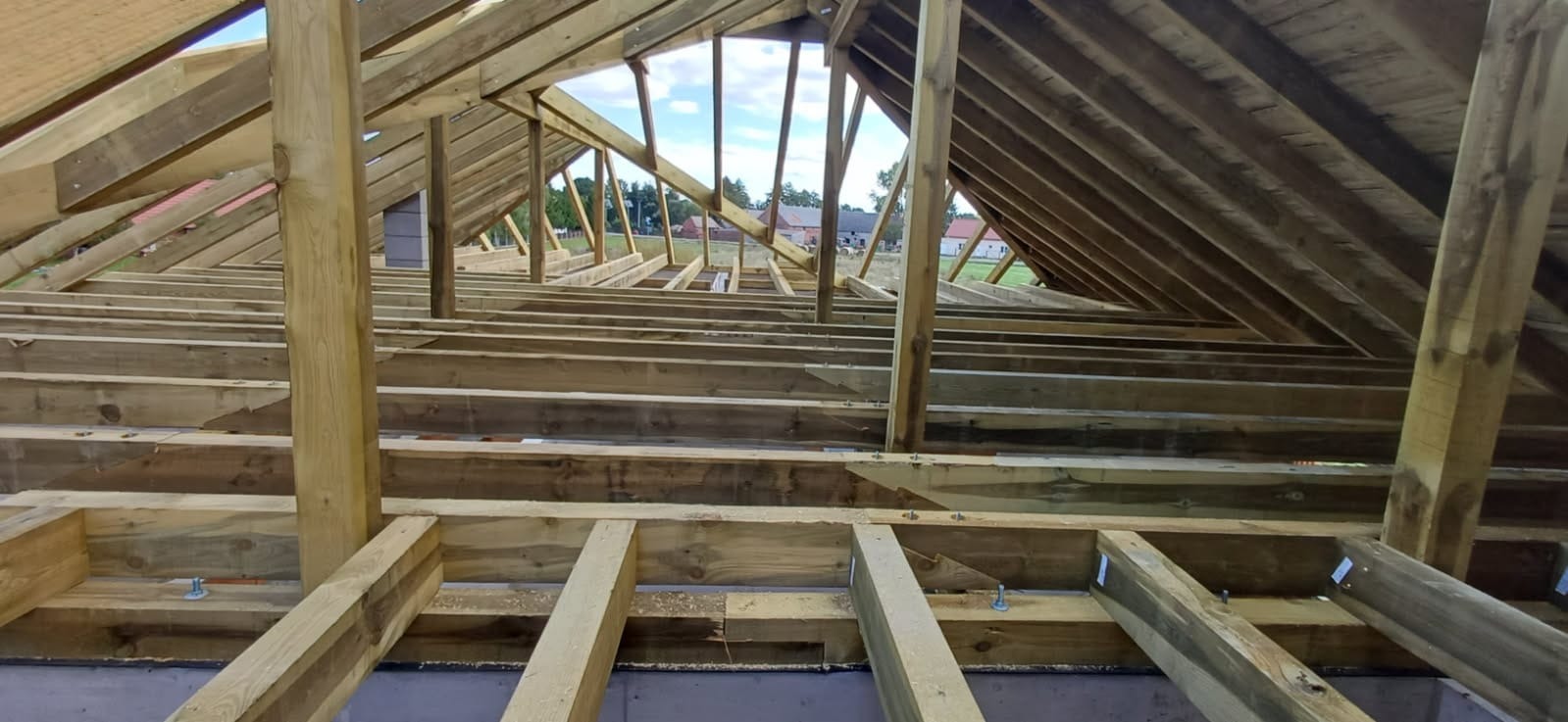 Konstrukcja drewniana dachu w trakcie budowy, widoczne krokwie i belki stropowe, jasne drewno, elementy złączne. W tle zabudowa jednorodzinna i błękitne niebo.