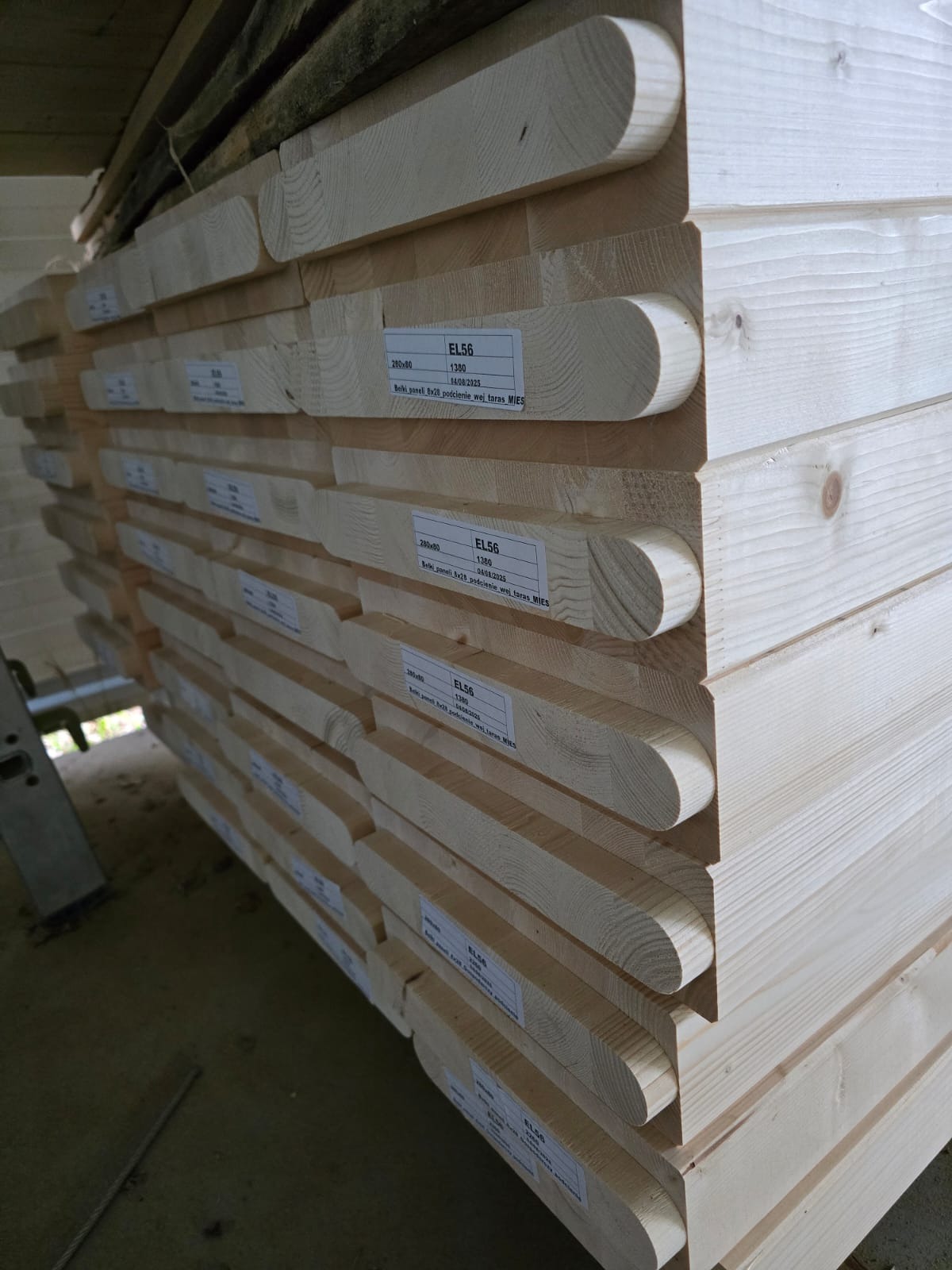 Stos jasnych belek drewnianych EL56 280x80 z etykietami, ułożonych równo. Widoczne słoje drewna i zaokrąglone krawędzie. Belki przeznaczone na podcienie, wejścia, tarasy.