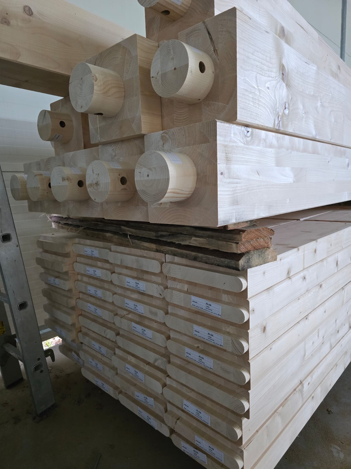 Stos tarcicy budowlanej z etykietami i drewnianych belek z okrągłymi czopami, ułożonych warstwami. Widoczna drabina po lewej stronie kadru.