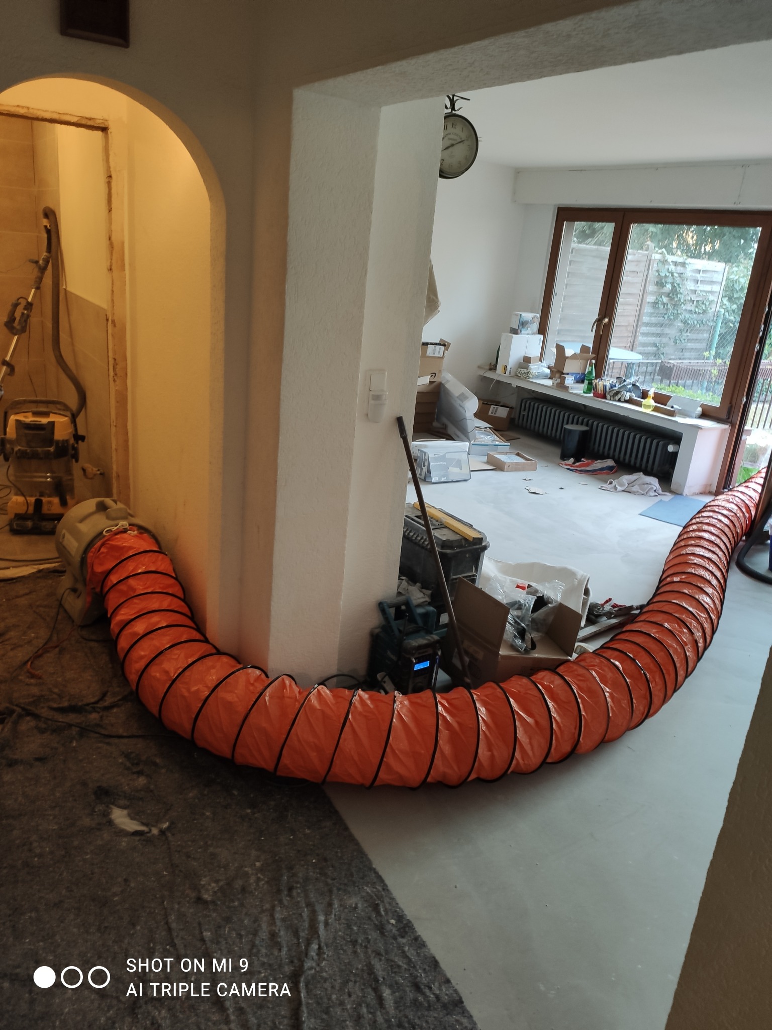 Pomarańczowy przewód wentylacyjny rozciągnięty przez remontowane pomieszczenie z surową podłogą, widoczne narzędzia i kartony, w tle okno i grzejnik.