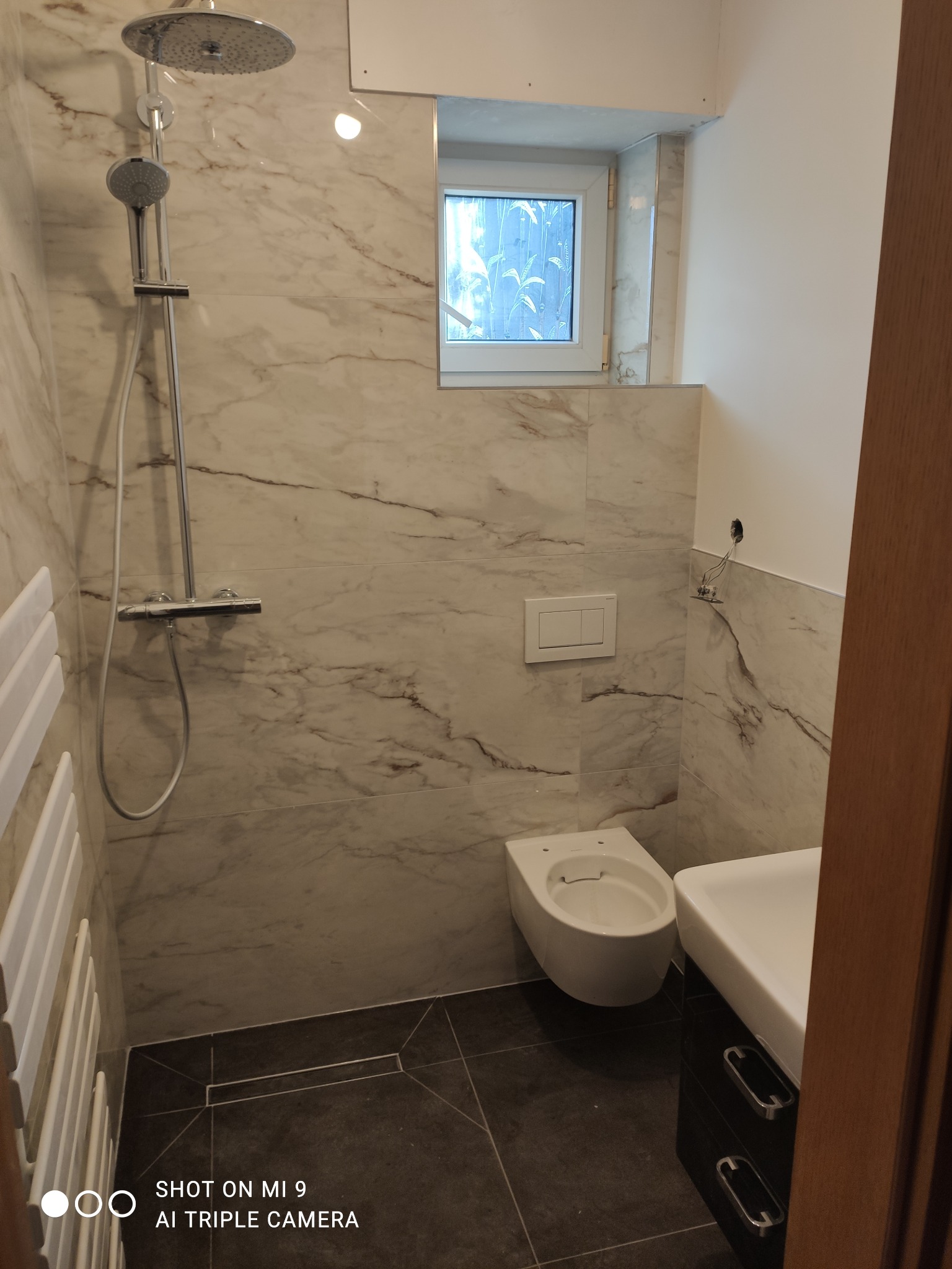 Nowoczesna łazienka z marmurowymi płytkami, kabiną prysznicową typu deszczownica, wiszącą miską WC i umywalką z szafką w kolorze czarnym, okno z widokiem na zieleń.