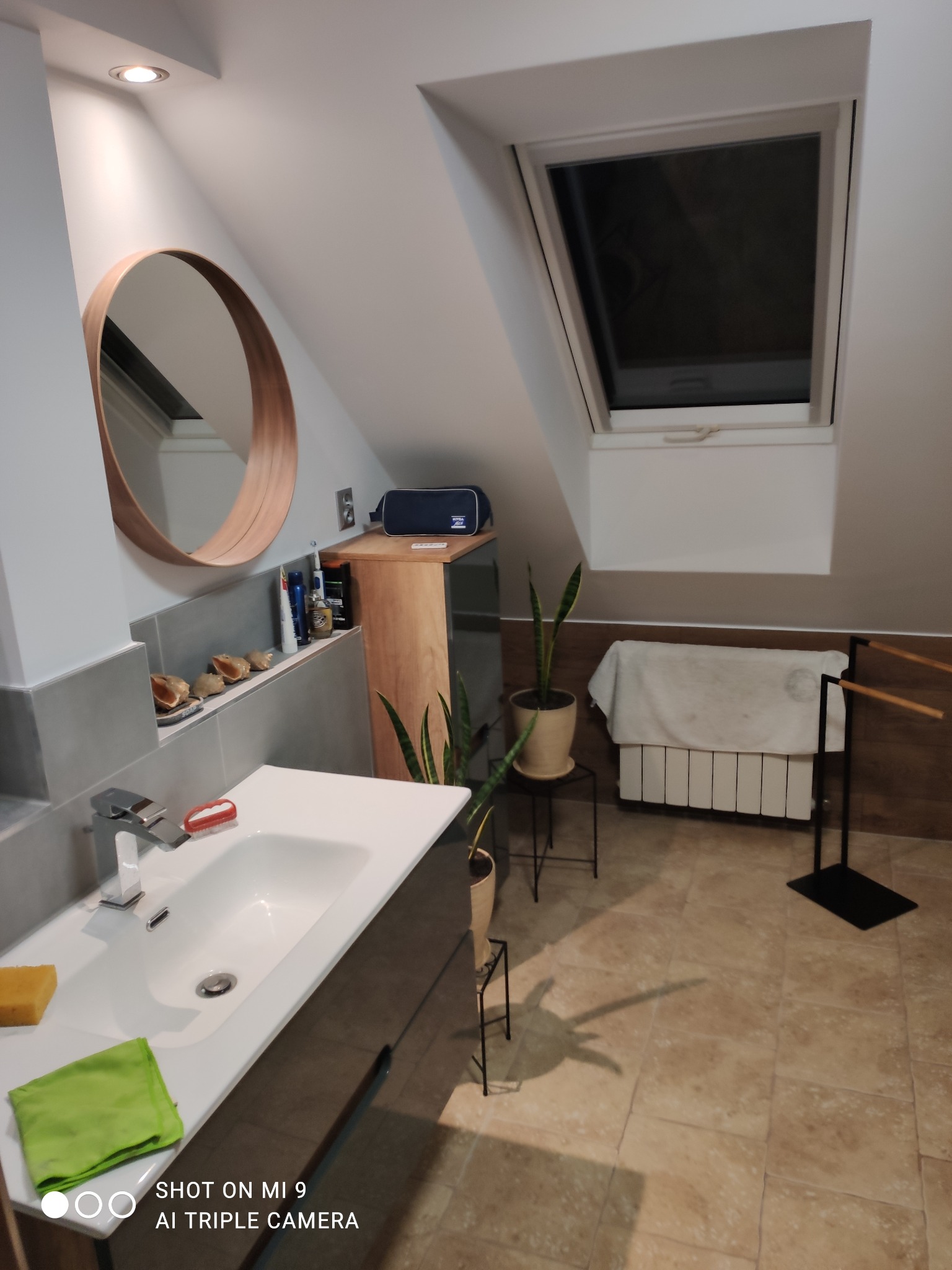 Nowoczesna łazienka na poddaszu z oknem dachowym, okrągłym lustrem z drewnianą ramą, białą umywalką i beżowymi płytkami podłogowymi. Na półce nad umywalką ustawione muszelki. Widoczny grzejnik...