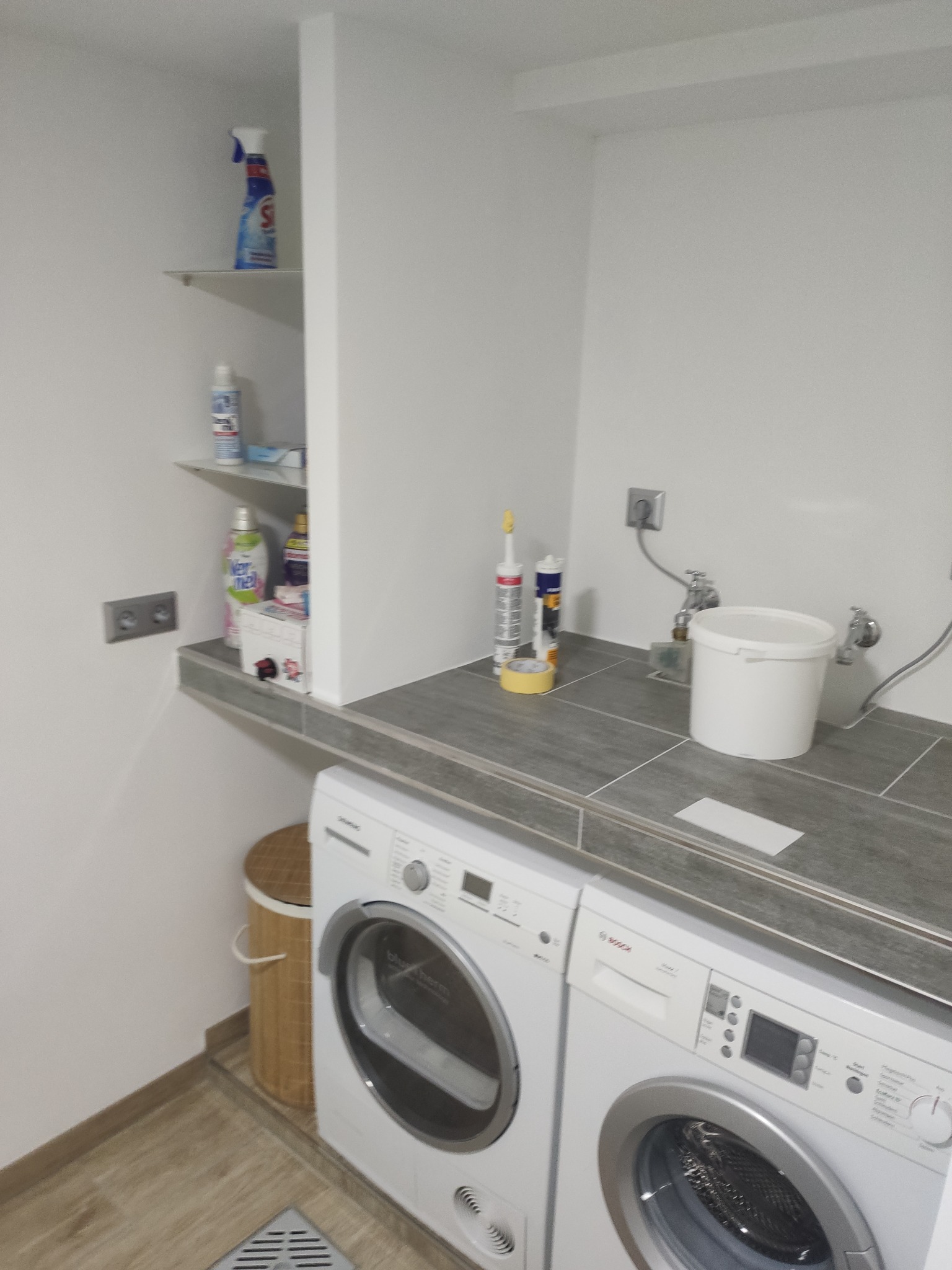 Szare płytki na ścianie i blacie w pralni, dwie białe pralki obok siebie, białe ściany i półki z detergentami, widoczne również akcesoria do prac wykończeniowych, takie jak silikony i taśma maskująca.