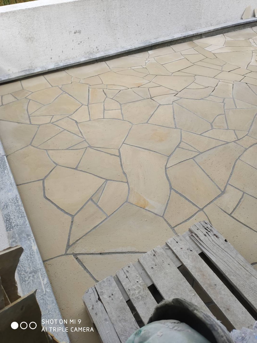 Ułożona mozaika z nieregularnych, piaskowych płytek na tarasie lub balkonie, widoczna paleta drewniana w rogu.